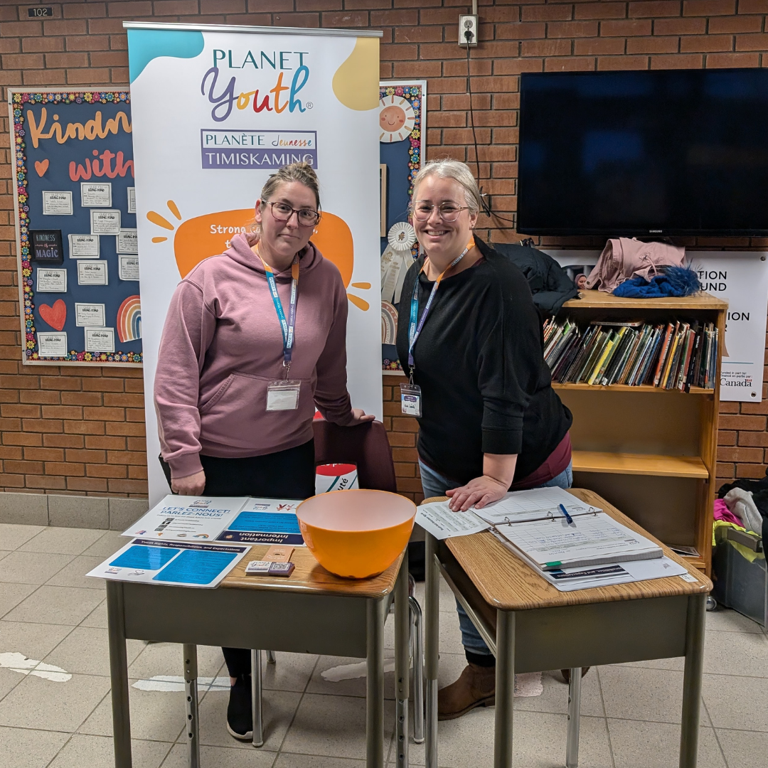 Two adult volunteers/ hub leads stand behind a table at a Planet Youth information booth inside a school hallway. The table is covered with pamphlets, forms, and a bowl. Behind them is a banner that reads "Planet Youth Timiskaming" with a slogan about supporting strong youth. The hallway is decorated with a bookshelf, bulletin boards, and school-related materials.