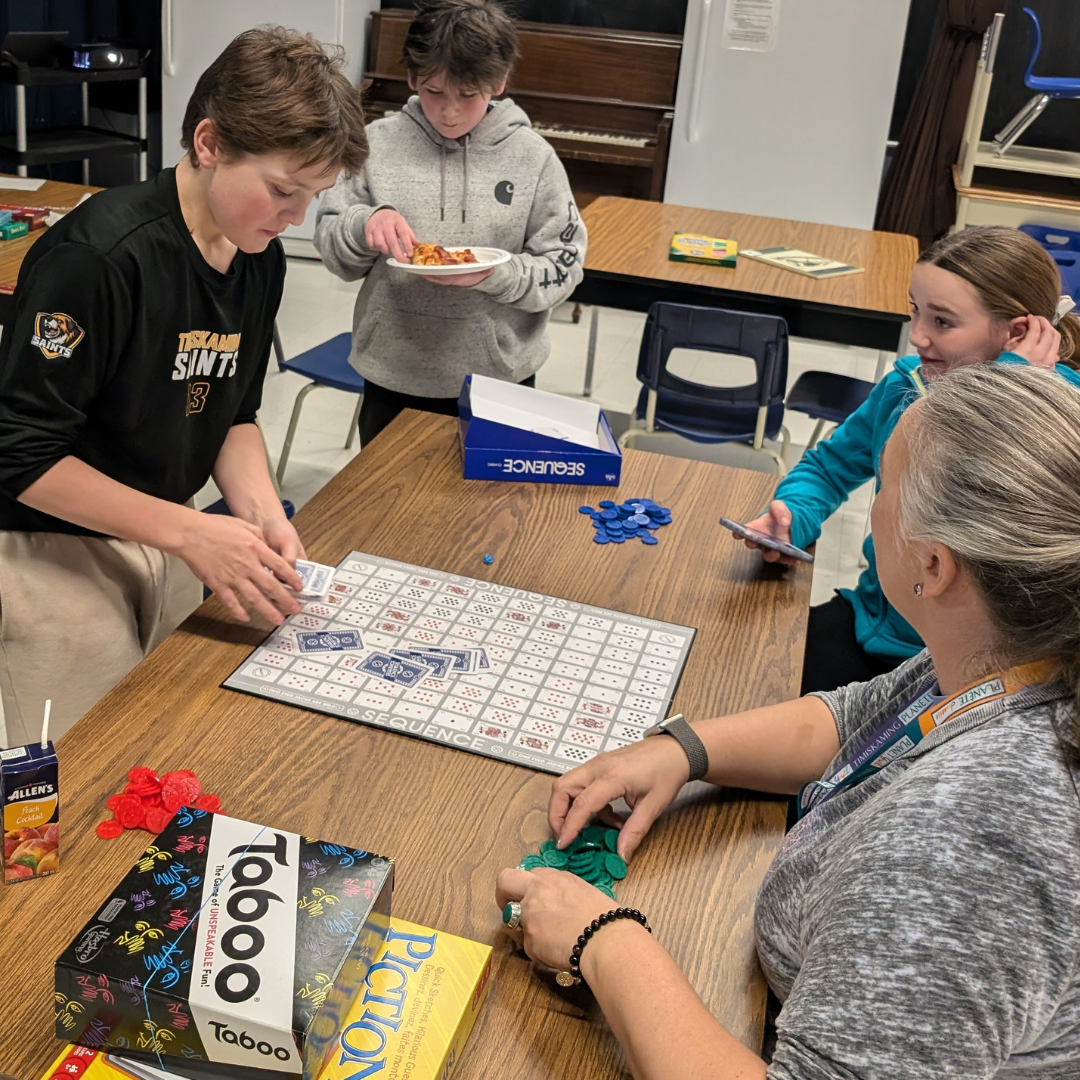 A group of young people and an adult (youth hub volunteer) are gathered around a table playing the board game "Sequence" at the youth hub. Other board games, including "Taboo" and "Pictionary," are visible on the table. A student in the background is eating pizza. The setting appears to be a classroom with multiple tables set up with other board games.