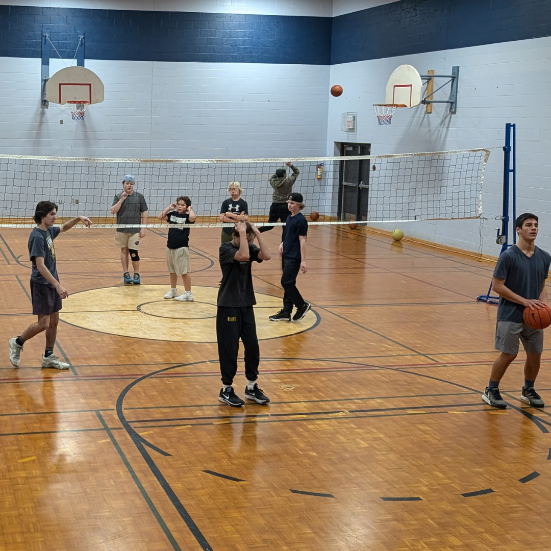 Youth are playing basketball and volleyball in a school gym. Some are holding basketballs while others are shooting hoops or practicing volleyball near the net.